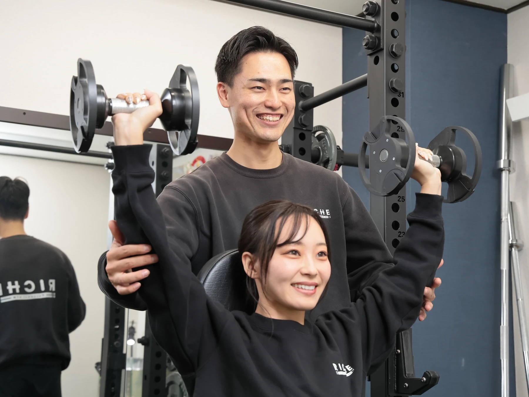 RICHEのジムで女性が男性トレーナーのサポートを受けてダンベルトレーニングに取り組む様子